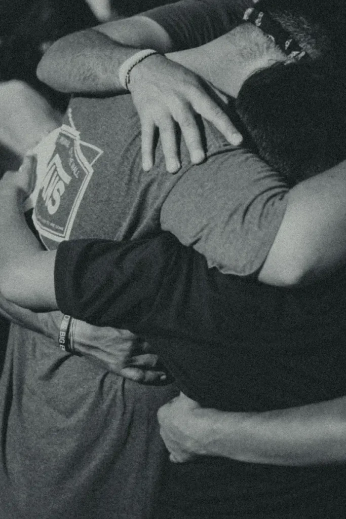 A close-up black-and-white photo of multiple people embracing in a group hug, showing hands and arms wrapped around each other in support and comfort.